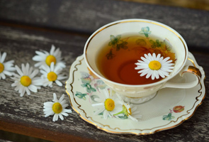 A floral-patterned teacup filled with tea sits on a matching saucer, with a chamomile flower floating in the tea and several daisies placed nearby on a wooden surface.